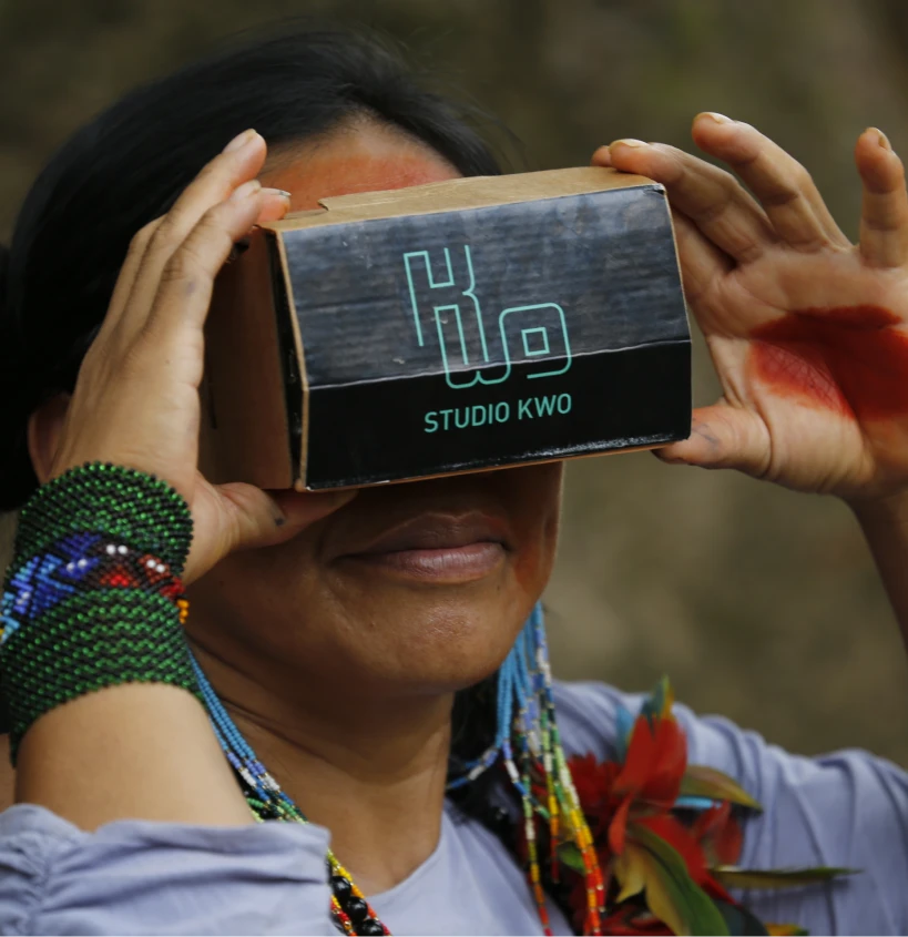 Close-up de mulher indígena sorrindo enquanto utiliza um óculos de realidade virtual com ologo do Estúdio Kwo